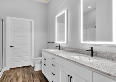 A white bathroom with a double vanity, lighted mirrors, and a glass shower.