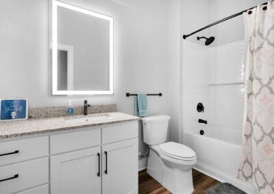 A white bathroom with a lighted mirror and a shower/tub combo with a shower curtain. A sign on the light quartz counter says "Please Seat Yourself" with a picture of a toilet.