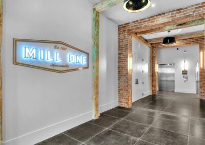 A hallway in the Mill One apartment building with decoratively weathered wooden arches and brick arches. A lighted sign reads "Mill One Mooresville, Est. 2021". There are at least two elevators at the end of the hall.