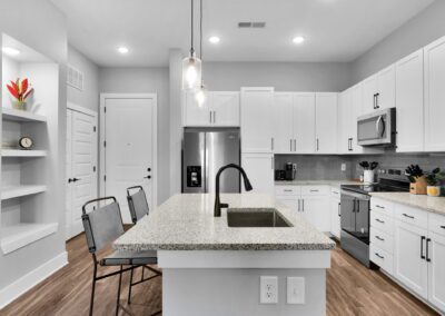 A clean white kitchen with stainless steel appliances and a spacious island in the center. There are built-in shelves behind the island.