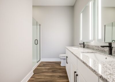 A white bathroom with a double vanity, lighted mirrors, and a large glass-door shower.